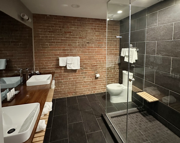Bathroom with brick accent wall, two white vessel sinks on a wooden counter, and a glass-enclosed shower. Towels are neatly hung and stored nearby.