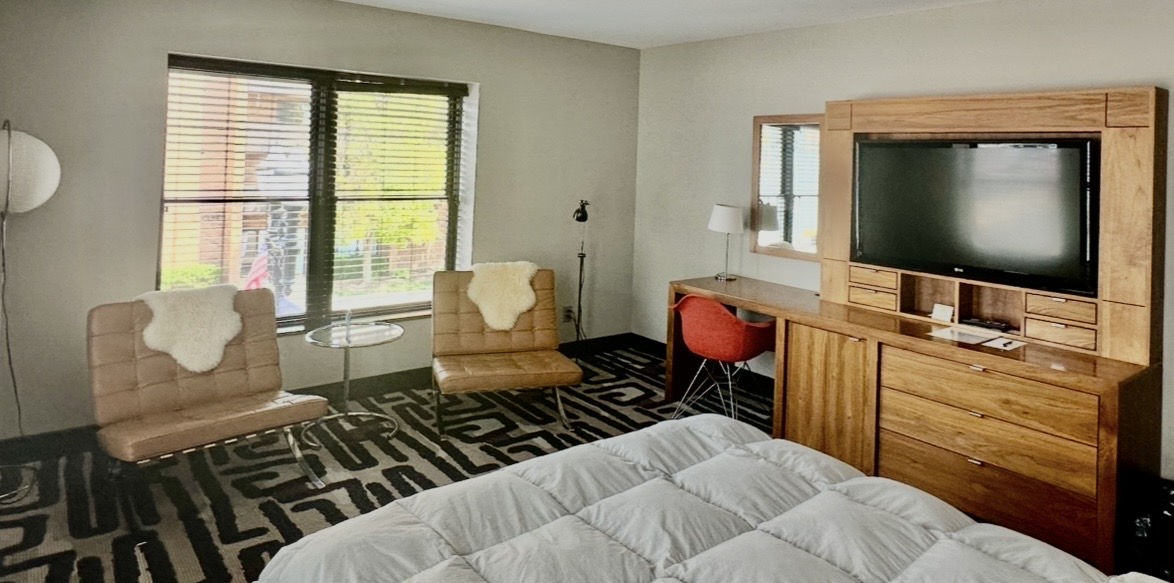 Hotel room with two tan lounge chairs near a window, a glass side table, and a large wooden TV cabinet in the background, bed in foreground.