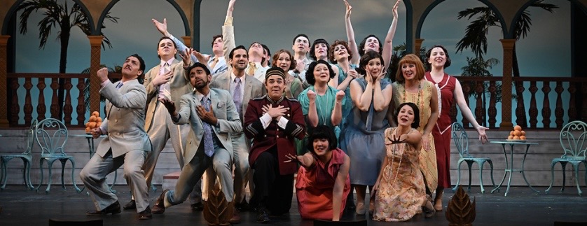 Large ensemble of actors on a vintage-styled stage, arms raised in a cheer, wearing colorful period costumes.