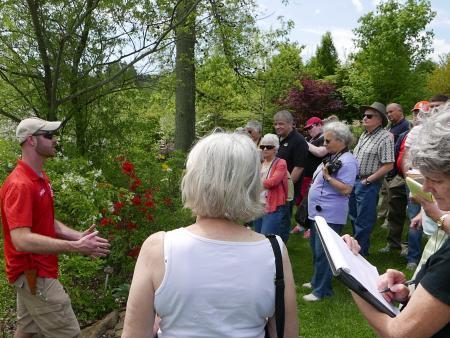 Tour guide in red shirt speaks to a group in a garden, with red flowers and greenery behind them.