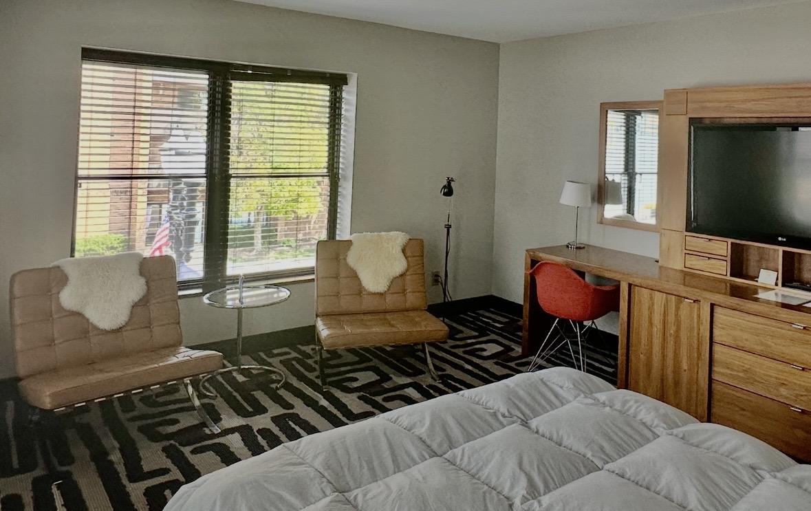Hotel room with two tan chairs by a large window, glass side table, and a bed in the foreground; wood desk, red chair, lamp, and flat-screen TV on a wooden console.