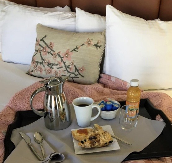 Breakfast tray on a bed with a coffee pot, white mug, pastry, juice bottle, and small yogurt cups.