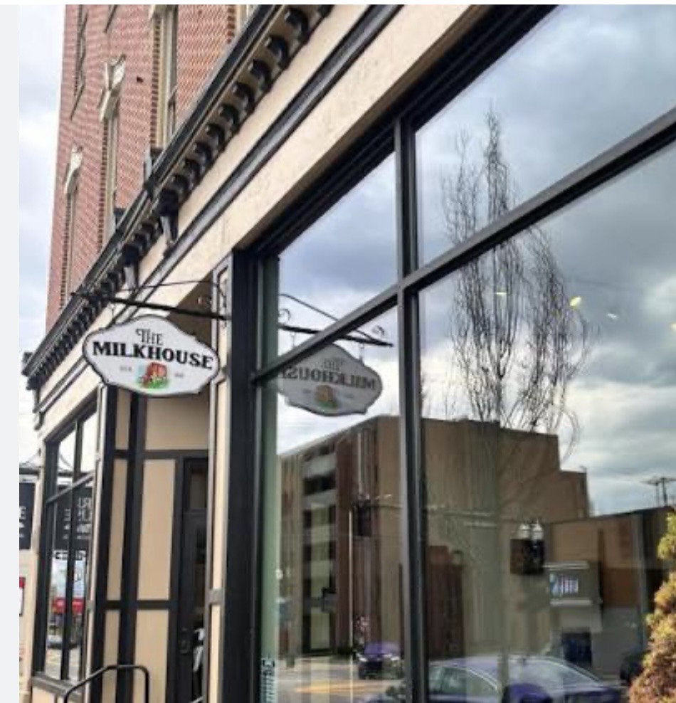Exterior of a storefront with a hanging sign reading 'The Milkhouse' beside large glass windows on a brick building.