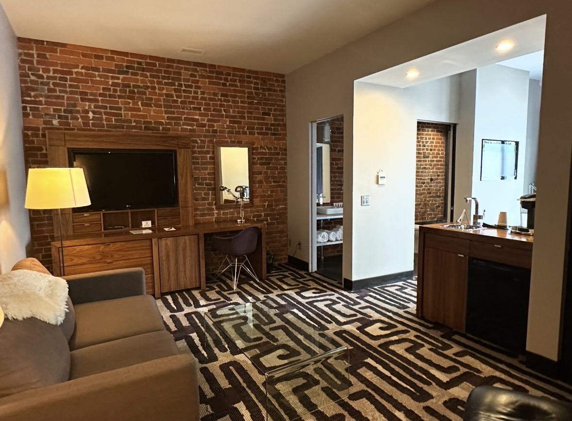 - St. Paul Hotel | Boutique Hotel Wooster Ohio Open-plan hotel living area with a brick accent wall, TV on a wooden console, sofa, and a glass coffee table beside a desk with a chair and mirror.
